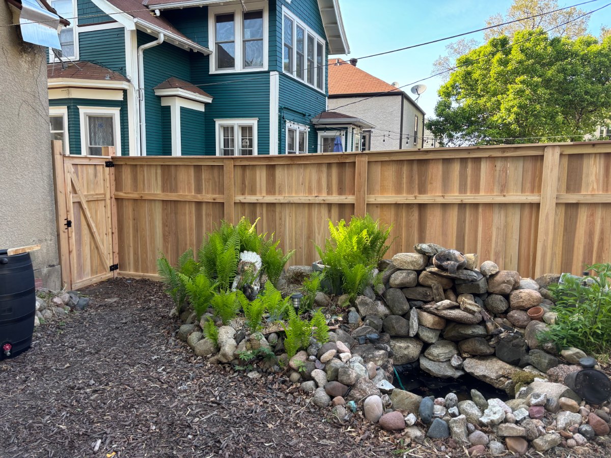 Stained cedar fence behind a rock garden and water feature, with a blue house beyond
